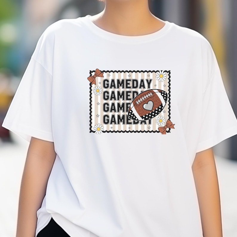 May include: White t-shirt with a football-themed graphic. The design features the word "GAMEDAY" repeated, a football with a heart, daisies, and brown bows. The shirt is a casual, everyday wear item.