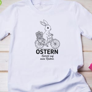 K&ouml;nnte beinhalten: Wei&szlig;es T-Shirt mit einer schwarz-wei&szlig;en Grafik eines Hasen, der Fahrrad f&auml;hrt und einen Korb mit Ostereiern hat. Der Text "OSTERN Kommt auf zwei R&auml;dern" ist unter dem Fahrrad aufgedruckt.