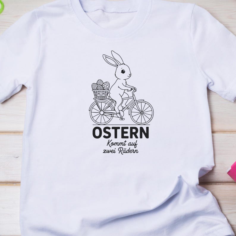 K&ouml;nnte beinhalten: Wei&szlig;es T-Shirt mit einer schwarz-wei&szlig;en Grafik eines Hasen, der Fahrrad f&auml;hrt und einen Korb mit Ostereiern hat. Der Text "OSTERN Kommt auf zwei R&auml;dern" ist unter dem Fahrrad aufgedruckt.