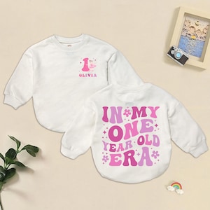 May include: White long-sleeved sweatshirt with pink text and graphics. The front of the shirt has the number 1 and the name "Olivia". The back of the shirt says "IN MY ONE YEAR OLD ERA" in pink and purple.