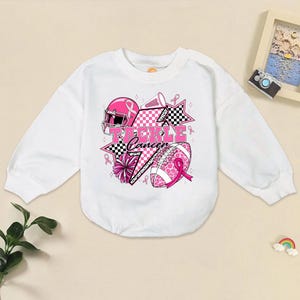 May include: White long-sleeve sweatshirt with a pink and black graphic. The graphic includes a football helmet, football, stars, and the words "Tackle Cancer". Pink ribbons and a pom-pom complete the design.