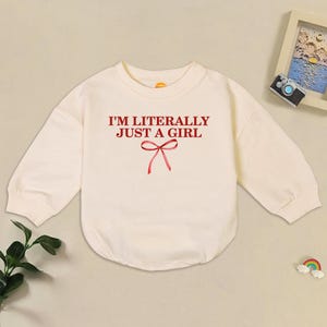 May include: Cream-colored baby bodysuit with long sleeves. The front features the text "I'M LITERALLY JUST A GIRL" in burgundy, with a red bow graphic underneath. The garment is made of soft fabric.
