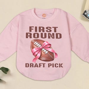 May include: A light pink sweatshirt with long sleeves. The front features a brown football tied with a pink bow, and the words "FIRST ROUND" and "DRAFT PICK" in brown lettering.