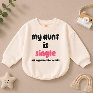 May include: A cream-colored long-sleeve sweatshirt featuring the black text "my aunt" and "is" with the pink text "single" and the black text "ask my parents for details" printed on the front. The garment has a crew neckline and ribbed cuffs.