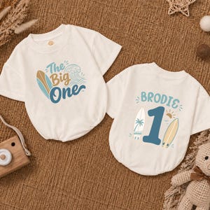 May include: Two white baby bodysuits with surf-themed graphics. One says "The Big One" with a surfboard and wave design. The other says "Brodie 1" with a surfboard, palm tree, and sun graphic. Both have short sleeves.