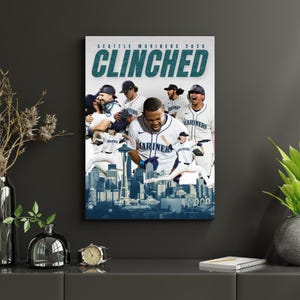 May include: A framed print featuring the Seattle Mariners baseball team celebrating a victory. The word "CLINCHED" is prominently displayed in teal above a collage of players in white and navy uniforms. The Seattle skyline is visible in the background.