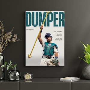 Pode incluir: Impressão emoldurada com um jogador de beisebol segurando um tridente dourado. A palavra "DUMPER" é exibida em letras grandes turquesas. O texto inclui "BIG DUMPER", "CAL RALEIGH", "SEATTLE MARINERS" e "HOMERUN DERBY 2025".