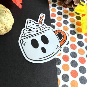 May include: A white ghost-shaped mug with a black outline and a red and white striped straw. The mug is filled with white whipped cream with black stars and white dots. The mug is on a black and white polka dot background.