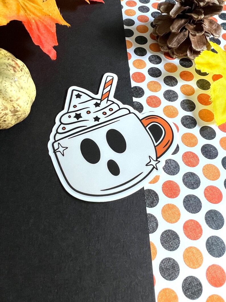 May include: A white ghost-shaped mug with a black outline and a red and white striped straw. The mug is filled with white whipped cream with black stars and white dots. The mug is on a black and white polka dot background.