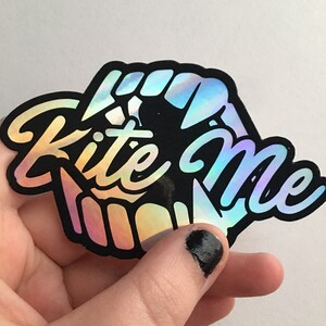 May include: A black and iridescent sticker with the text "Kite Me" in a stylized font. The sticker features a kite design with a rainbow iridescent finish.