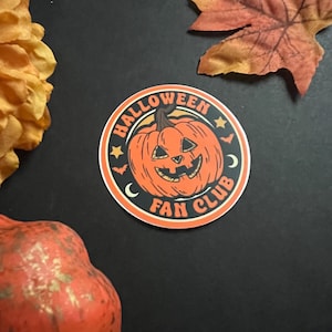 Pu&ograve; includere: Un adesivo rotondo con una zucca arancione con un viso di jack-o'-lantern. L'adesivo dice "Halloween Fan Club" in un cerchio nero con stelle bianche e mezze lune.