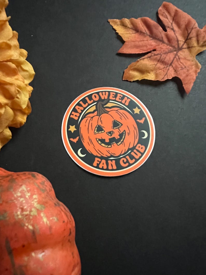 Pu&ograve; includere: Un adesivo rotondo con una zucca arancione con un viso di jack-o'-lantern. L'adesivo dice "Halloween Fan Club" in un cerchio nero con stelle bianche e mezze lune.