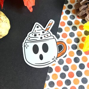 May include: A white ghost-shaped coffee cup with a black and white whipped cream topping and a red and white straw. The cup has a black outline and a black handle. The background is a black and white polka dot pattern.