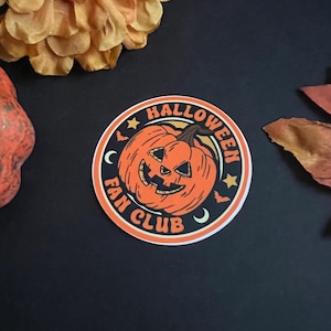 Pu&ograve; includere: Un adesivo rotondo con una zucca arancione con un viso spettrale, una luna crescente e stelle. Il testo "HALLOWEEN FAN CLUB" &egrave; scritto intorno alla zucca.