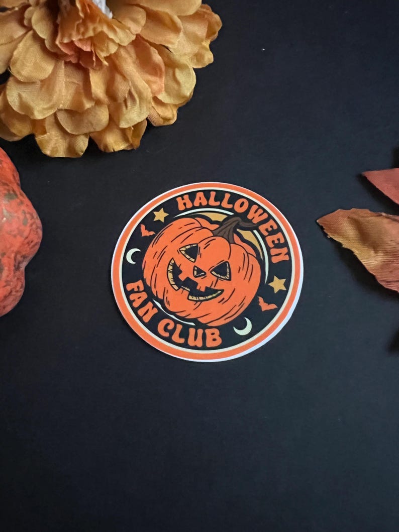 Pu&ograve; includere: Un adesivo rotondo con una zucca arancione con un viso spettrale, una luna crescente e stelle. Il testo "HALLOWEEN FAN CLUB" &egrave; scritto intorno alla zucca.