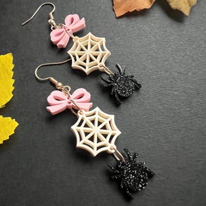 May include: A pair of Halloween-themed earrings. Each earring features a pink bow, a cream-colored spider web, and a black glittery spider. The earrings are suspended from silver-colored hooks.