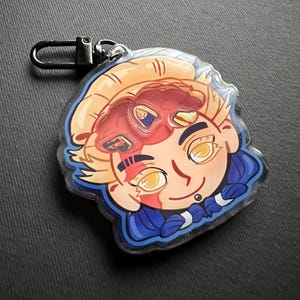 May include: A cartoon-style keychain featuring a character with blonde hair, yellow eyes, and a blue outfit. The character has a red mask and a small piercing. The keychain has a black clasp.