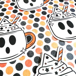 May include: Four white ghost-shaped mugs with black eyes and mouths, filled with white whipped cream and topped with a red and white striped straw and black stars. The mugs are on a background of orange and black polka dots.