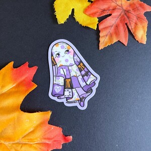 Op de afbeelding: Een sticker met een cartoongeest met een patchwork-ontwerp in paars, wit en oranje. De geest heeft een lachend gezicht met bloemaccenten. De sticker is omgeven door herfstbladeren en een kleine pompoen.