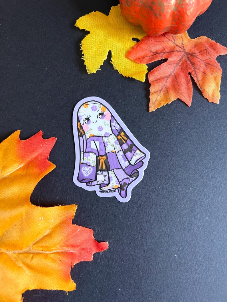 Op de afbeelding: Een sticker met een cartoongeest met een patchwork-ontwerp in paars, wit en oranje. De geest heeft een lachend gezicht met bloemaccenten. De sticker is omgeven door herfstbladeren en een kleine pompoen.