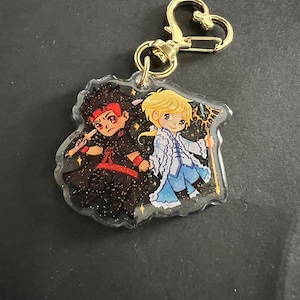May include: A clear acrylic keychain featuring two anime characters. The character on the left is wearing a black and red outfit and holding a weapon. The character on the right is wearing a blue and white outfit and holding a staff. The background is a sparkly gold.