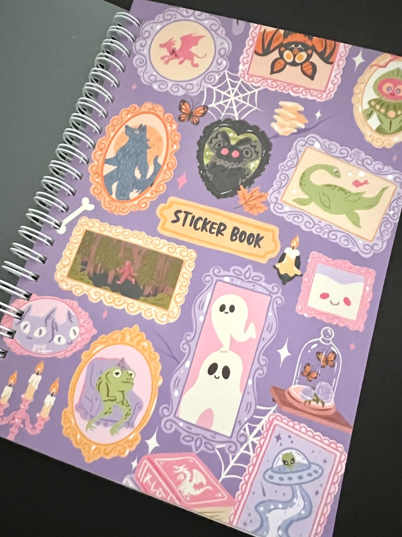 May include: A spiral-bound sticker book with a lilac cover. The cover is decorated with framed illustrations of a wolf, a frog, ghosts, and a dragon. The words "Sticker Book" are printed on a yellow banner. The illustrations have a whimsical style.