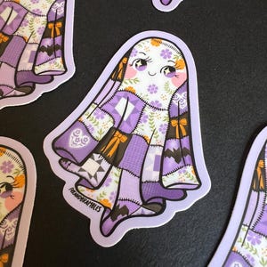 Op de afbeelding: Een verzameling stickers met een vriendelijk spookontwerp. Het spook heeft een patchwork-stijl lichaam in tinten paars, wit en bruin, met bloemen- en strikaccenten. De stickers hebben een lichtpaarse rand en een lachend gezicht.