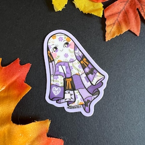 May include: A whimsical sticker featuring a smiling ghost with a patchwork design in shades of purple, white, and orange. The ghost has a floral pattern on its face and body, with a bow detail. The sticker is surrounded by colorful autumn leaves.