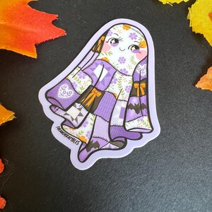 Op de afbeelding: Een sticker met een vriendelijk spook met een patchwork quilt ontwerp in tinten paars, wit en bruin. Het spook heeft een lachend gezicht met roze wangen en bloemaccenten. De sticker heeft de tekst "FOXANDBRAMBLES" linksonder.
