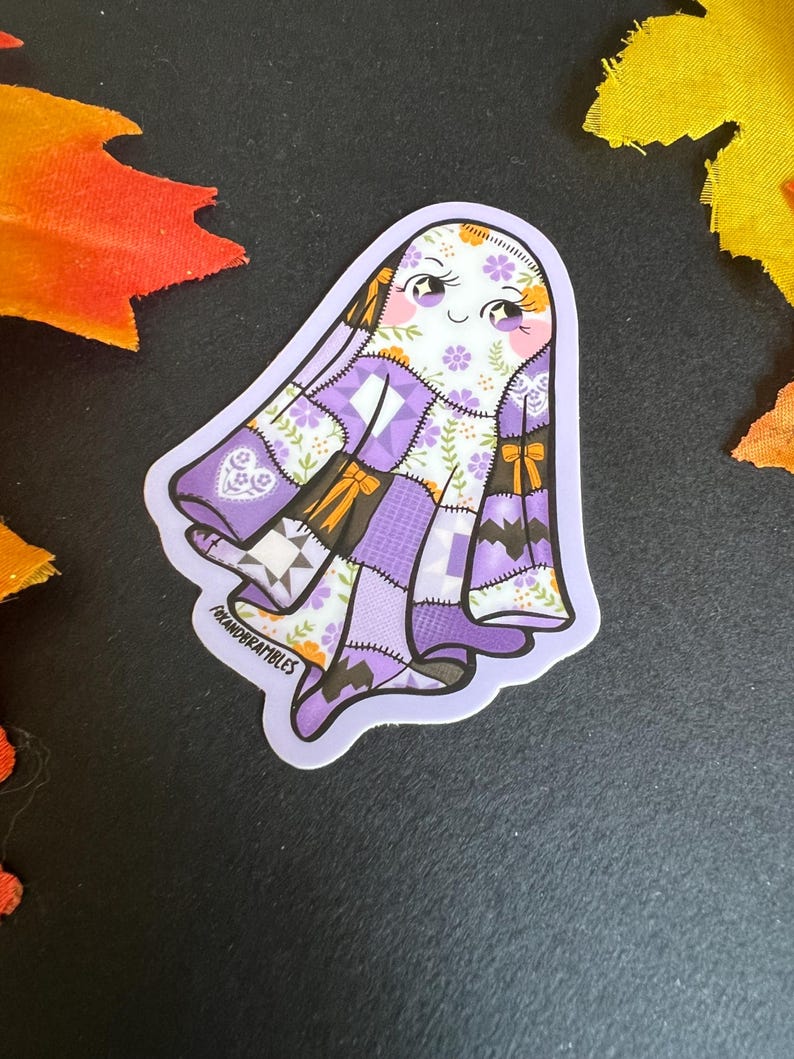 Op de afbeelding: Een sticker met een vriendelijk spook met een patchwork quilt ontwerp in tinten paars, wit en bruin. Het spook heeft een lachend gezicht met roze wangen en bloemaccenten. De sticker heeft de tekst "FOXANDBRAMBLES" linksonder.