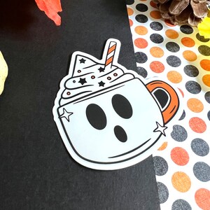 May include: A white ghost-shaped mug with a red and white striped straw, filled with white whipped cream and black stars. The mug has a black outline and a black handle.