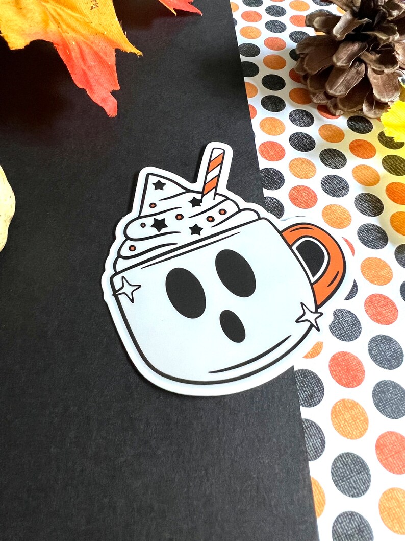 May include: A white ghost-shaped mug with a red and white striped straw, filled with white whipped cream and black stars. The mug has a black outline and a black handle.