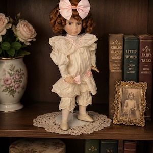 Puede incluir: Una muñeca de porcelana con cabello castaño rojizo y un gran lazo rosa, vestida con un vestido color crema con adornos de encaje y un lazo a juego. La muñeca está sobre un tapete, con libros antiguos y una foto enmarcada en el fondo. El atuendo de la muñeca incluye un cuello con volantes y bombachos.