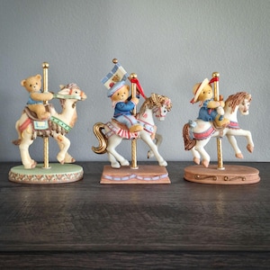 Może przedstawiać: Trzy figurki karuzeli z misiami jadącymi na wielbłądzie i dwóch koniach. Figurki są pomalowane pastelowymi kolorami ze złotymi akcentami. Każdy miś ubrany jest w inny strój, a zwierzęta z karuzeli mają szczegółowe siodła i dekoracje.