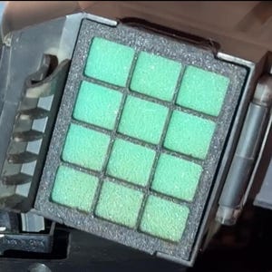 Puede incluir: Primer plano de un objeto rectangular con una cuadrícula de doce almohadillas cuadradas de espuma verde claro. Las almohadillas están colocadas en un marco negro, con una carcasa metálica gris oscuro que rodea el marco.