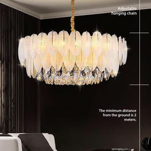 Może przedstawiać: Złoty żyrandol z pióropodobnym wzorem i kryształowymi akcentami. Tekst wskazuje na regulowany łańcuch do zawieszania i minimalną odległość 2 metrów od podłoża. Oprawa wisi nad białą sofą w ciemnym pomieszczeniu.