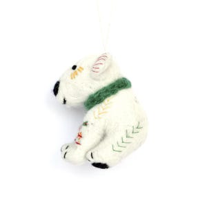 May include: A white felt dog ornament with a green collar and black accents. The dog has embroidered details in yellow, red, and green. A white string is attached for hanging.