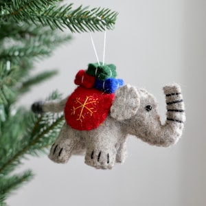 May include: A gray felt elephant Christmas ornament with a red saddle featuring a gold snowflake design. The elephant carries green, red, and blue bundles on its back. The ornament is hanging from a Christmas tree.