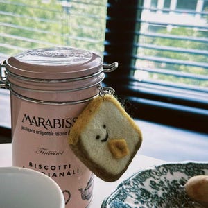 May include: A pink metal container with the text "MARABISS" and a felt bread slice keychain with a smiling face. The container is next to a plate with a floral design and a white teacup.