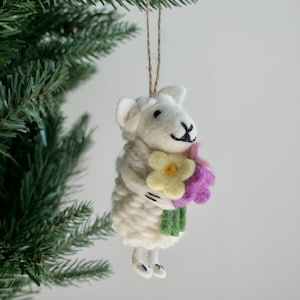 May include: A white sheep ornament with a smiling face and a bouquet of flowers. The sheep is holding a bouquet of yellow, purple, and green flowers. The ornament is hanging from a brown string, and is next to a green Christmas tree.