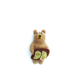 May include: A handmade brown bear figurine with a yellow snout and eyes, holding a brown object with two green spots. The bear is made of felt and stands upright against a white background.