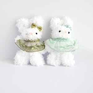 May include: Two fluffy white bunny plush toys with black eyes, pink noses, and decorative collars. One bunny has a green bow and collar, the other has a light green bow and collar. The bunnies are standing on a white surface.