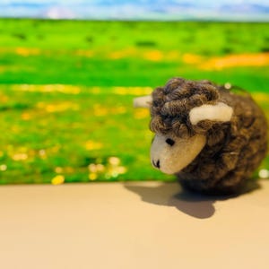 May include: A handcrafted, gray wool sheep figurine with white accents on the face and ears. The sheep is set against a blurred background of green and blue, suggesting a pastoral scene. The figurine is a charming decorative item.