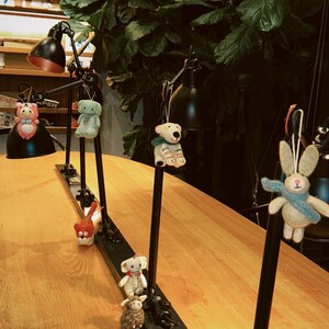 May include: A display of decorative animal ornaments hanging from black metal lamps. The ornaments include a pink bear, a green elephant, a red cat, a white bear, a white rabbit, a white elephant, and a brown sheep. The lamps are on a wooden table.