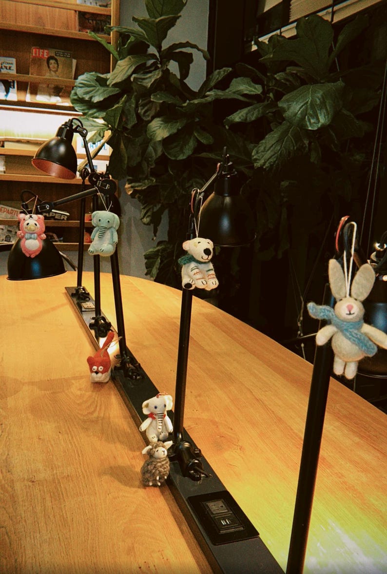 May include: A display of decorative animal ornaments hanging from black metal lamps. The ornaments include a pink bear, a green elephant, a red cat, a white bear, a white rabbit, a white elephant, and a brown sheep. The lamps are on a wooden table.