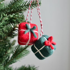 May include: Two Christmas ornaments shaped like wrapped gifts. One is red with a green bow, the other is green with a red bow. Both are decorated with white string and hang from red and white twine.