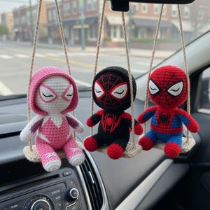 May include: Three crocheted superhero figures hanging from a car's rearview mirror. The figures include Spider-Gwen in pink, Miles Morales in black, and Spider-Man in red and blue, each seated on a small swing.