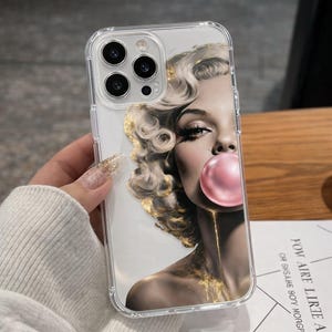 May include: Clear phone case featuring a portrait of a woman with blonde curly hair blowing a pink bubble. The image has gold accents and is set against a white background. The case has a clear border and a camera cutout.