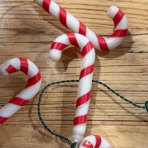 May include: A string of Christmas lights featuring white and red striped candy cane-shaped bulbs. The candy canes are arranged on a green wire against a wooden background, evoking a festive holiday theme.