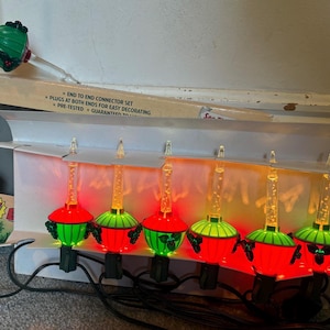 May include: A string of six bubble Christmas lights featuring red and green bases with holly leaf accents. Each light has a clear glass tube filled with liquid that bubbles when lit. The set is displayed on a cardboard backing with text.